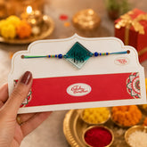 Rakhi with a card held by a hand, traditional setting with flowers and lights.