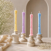 Four decorative candles in pastel colors on white candlesticks with a neutral background.