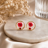 Red floral resin stud earrings with gold border displayed on a beige marble surface with soft luxury botanical background