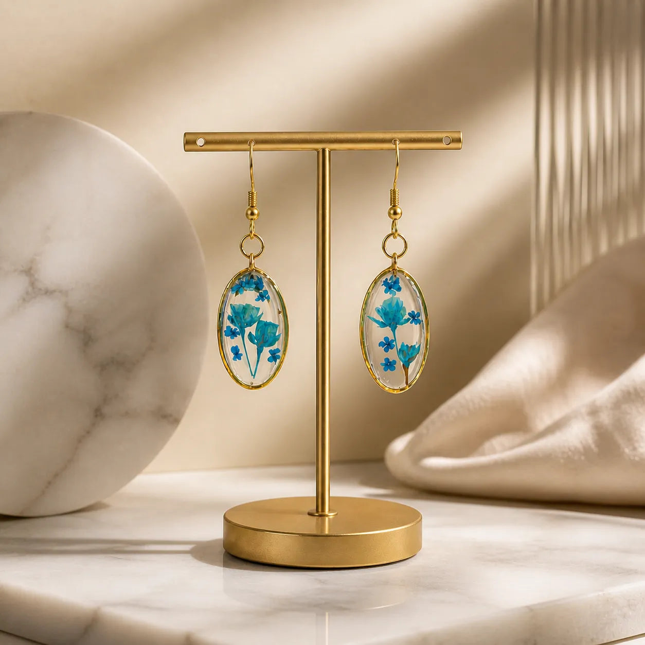 Blue floral resin oval drop earrings displayed on a gold stand with premium beige background, handmade lightweight jewellery for women