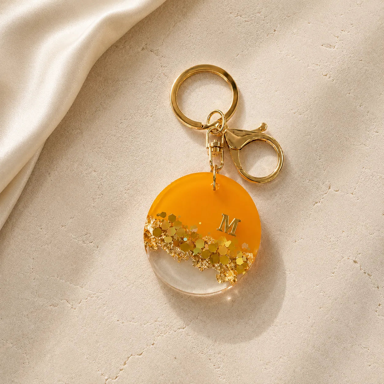 Gold keychain with a round orange pendant featuring gold glitter and the letter 'M' on a beige surface.
