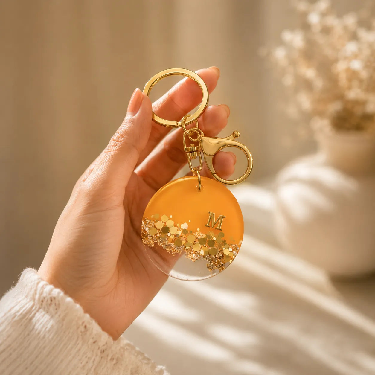 Hand holding a gold keychain with a yellow disc and letter 'M' against a neutral background