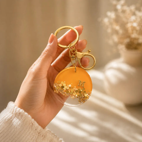 Hand holding a gold keychain with a yellow disc and letter 'M' against a neutral background