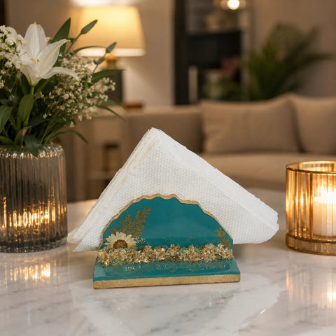 Decorative tissue holder with teal base and gold accents on a marble surface, with flowers and candle in the background.
