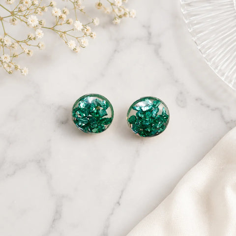 Top-view flat lay image of handmade emerald green crystal resin stud earrings placed on a white marble surface with soft luxury styling