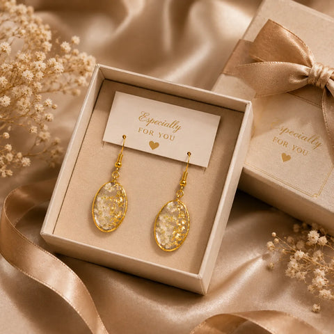 Floral resin oval drop earrings with white dried flowers and gold flakes displayed in a premium gift box setup with satin ribbon and elegant luxury background