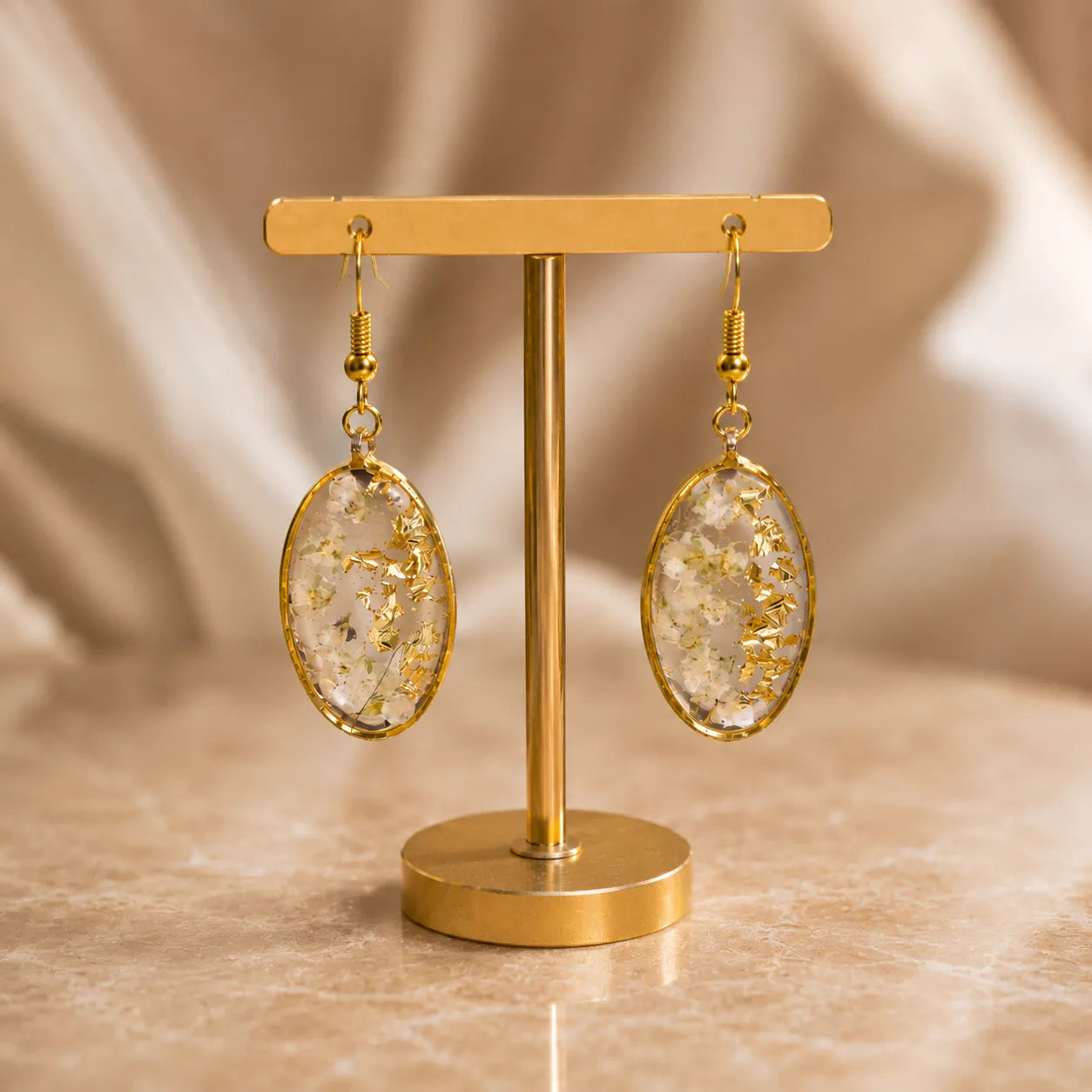 Floral resin oval drop earrings with white dried flowers, gold flakes, and gold border displayed on a premium golden stand with soft luxury background