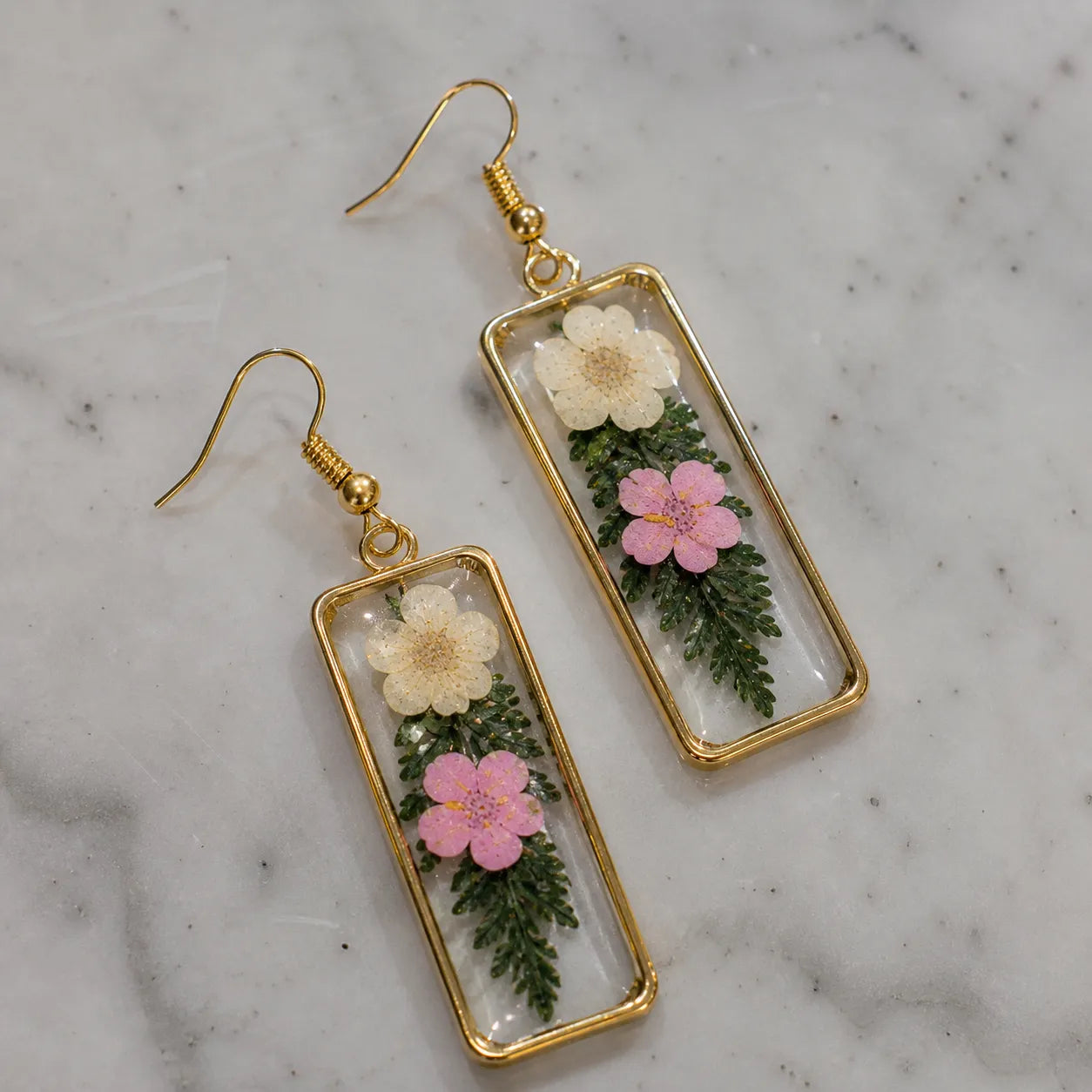 Floral resin rectangle drop earrings with white and pink dried flowers, transparent glossy finish, and elegant gold border placed on a marble surface