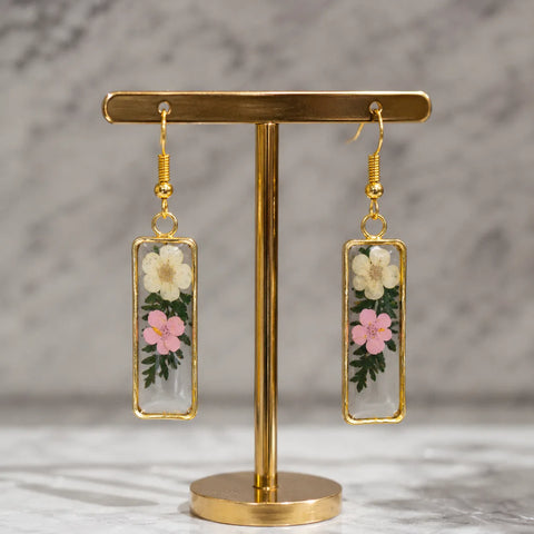 Floral resin rectangle drop earrings with white and pink dried flowers displayed on a premium golden stand with elegant marble background