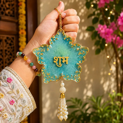 Hand holding a decorative blue star-shaped ornament with gold text and pearl tassel against a floral background.