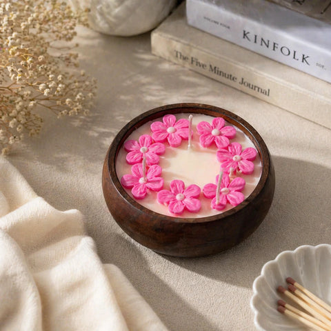Candle with pink floral designs in a wooden holder on a neutral surface with books and dried flowers.