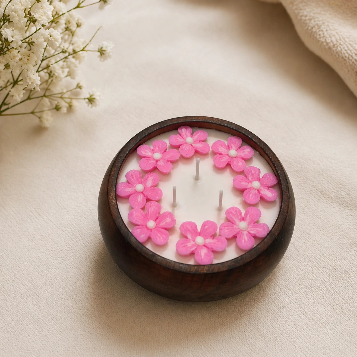 Candle with pink floral designs in a wooden holder on a beige surface with white flowers in the background.