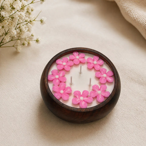 Candle with pink floral designs in a wooden holder on a beige surface with white flowers in the background.