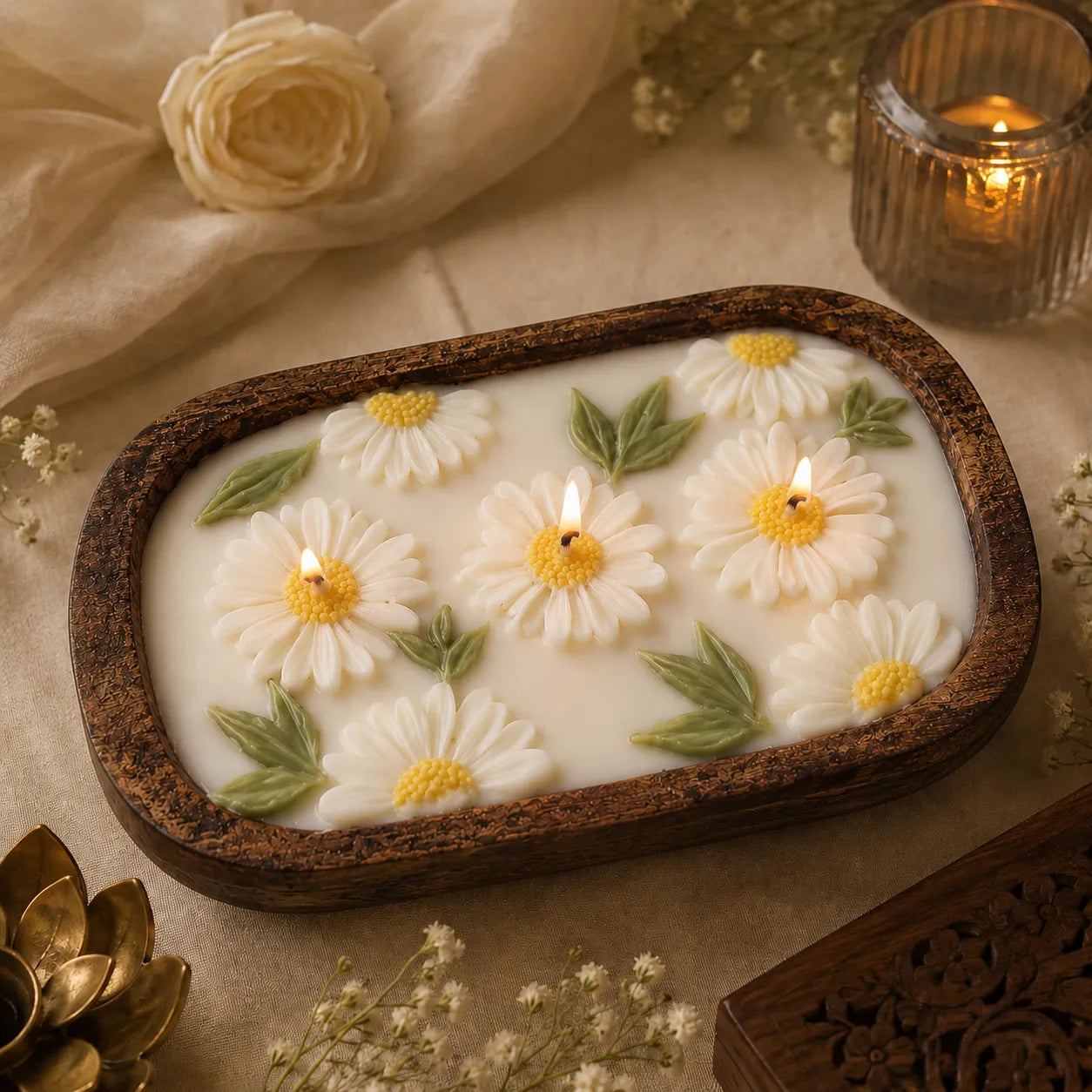 Candle with daisy flower designs on a wooden holder, surrounded by flowers and a candle.