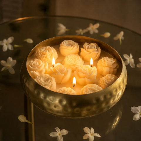 Decorative candle with rose-shaped wax pieces in a metallic bowl on a reflective surface with flowers.