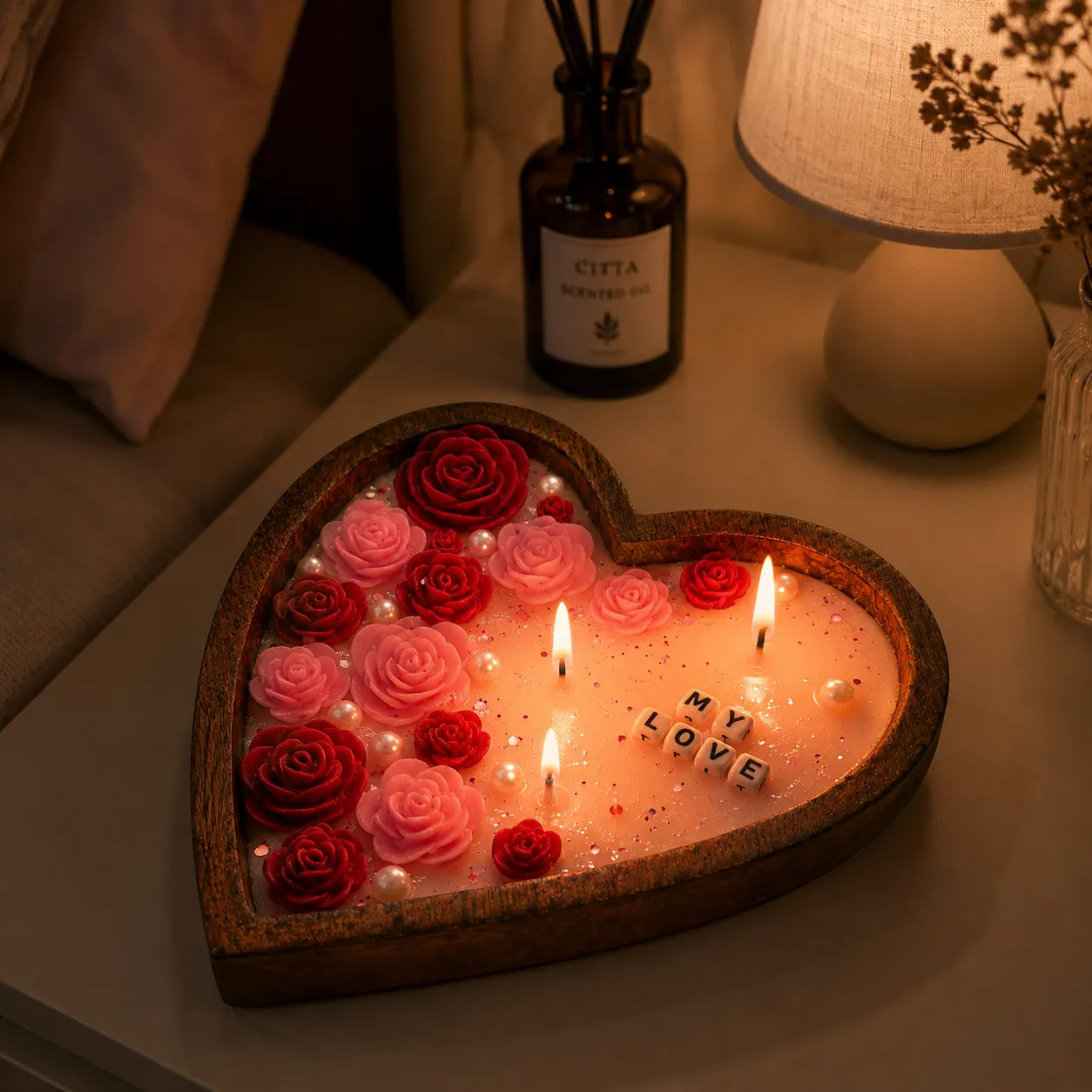 Heart-shaped candle with roses and 'LOVE' letters on a bedside table with a lamp and diffuser in the background.
