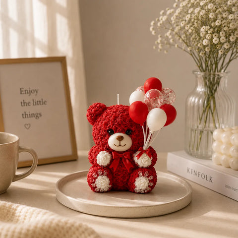 Red teddy bear candle with balloons on a wooden surface, next to a mug and a vase with flowers.