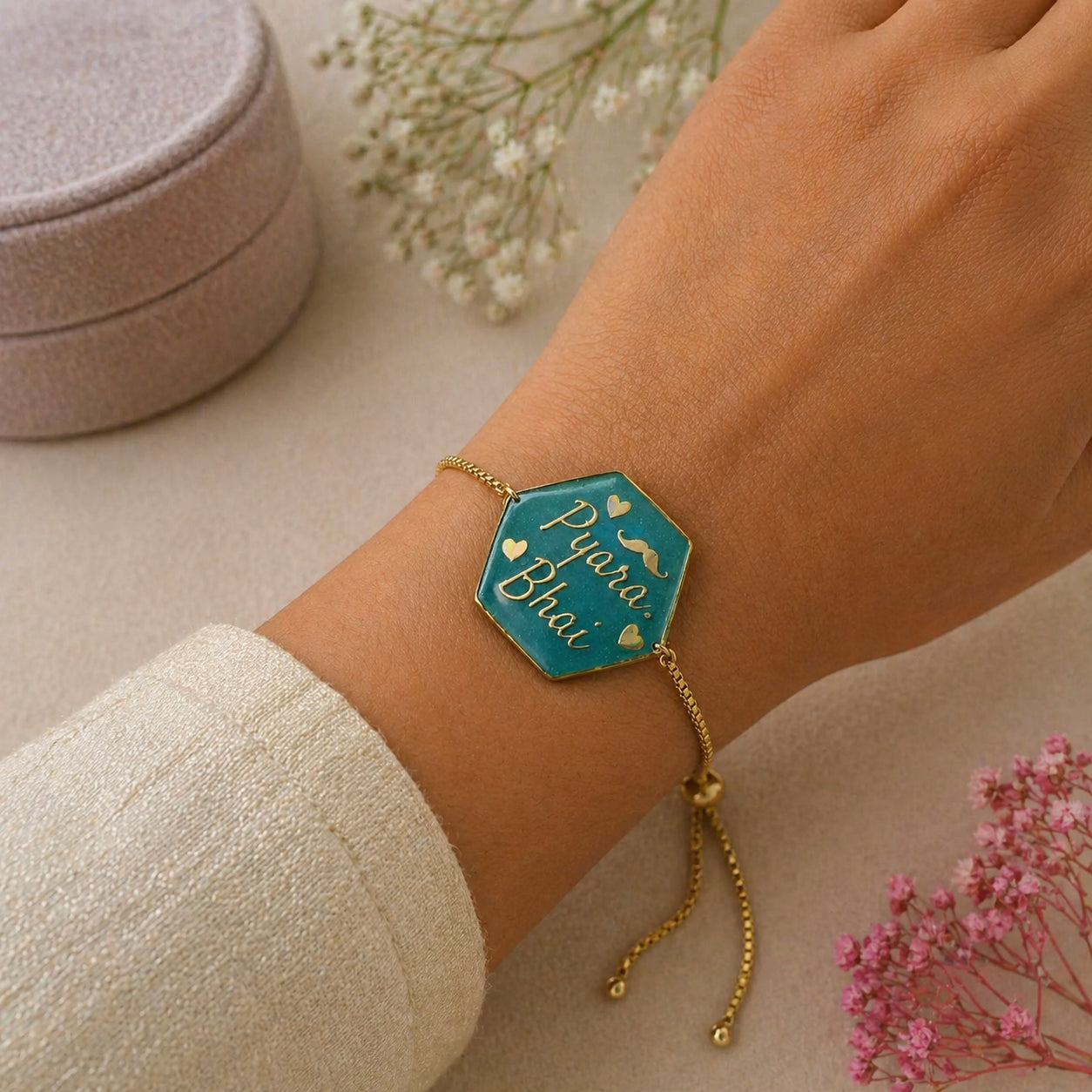 Bracelet with 'Pyara Bhai' on a wrist, with a pink jewelry box and flowers in the background.