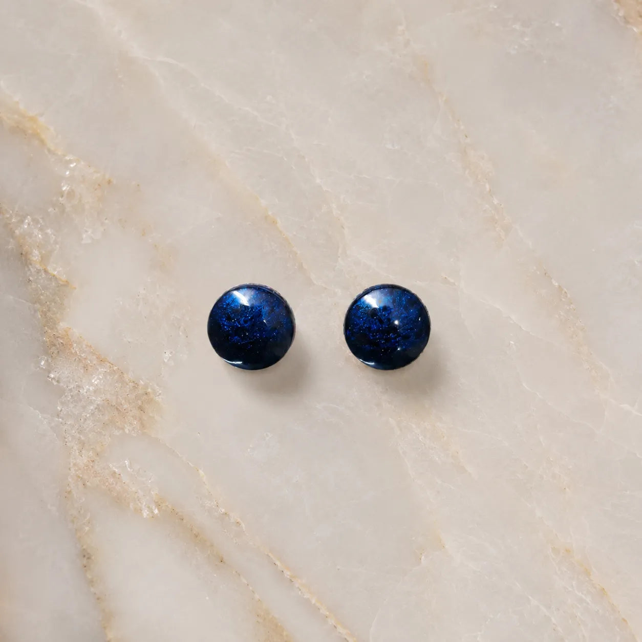Top-view flat lay image of handmade navy blue glossy resin stud earrings placed on a light marble surface with minimal luxury styling