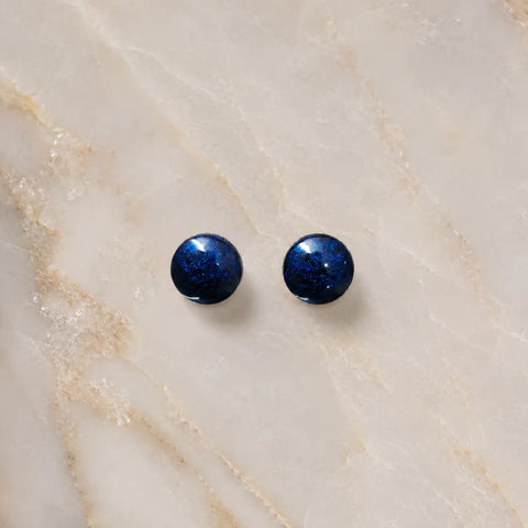 Top-view flat lay image of handmade navy blue glossy resin stud earrings placed on a light marble surface with minimal luxury styling