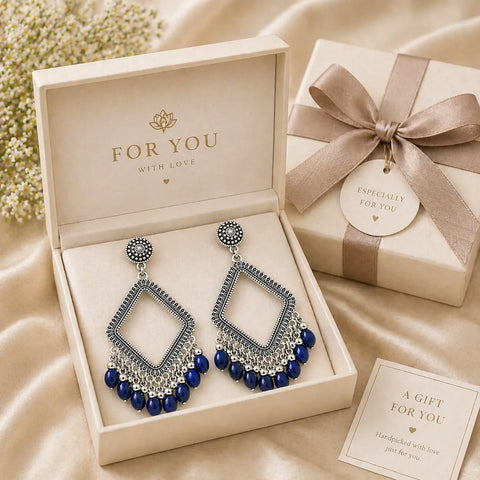 Oxidised silver diamond-shaped drop earrings with intricate detailing and blue bead fringe, elegantly placed inside a premium beige gift box with satin ribbon packaging on a soft neutral fabric background.