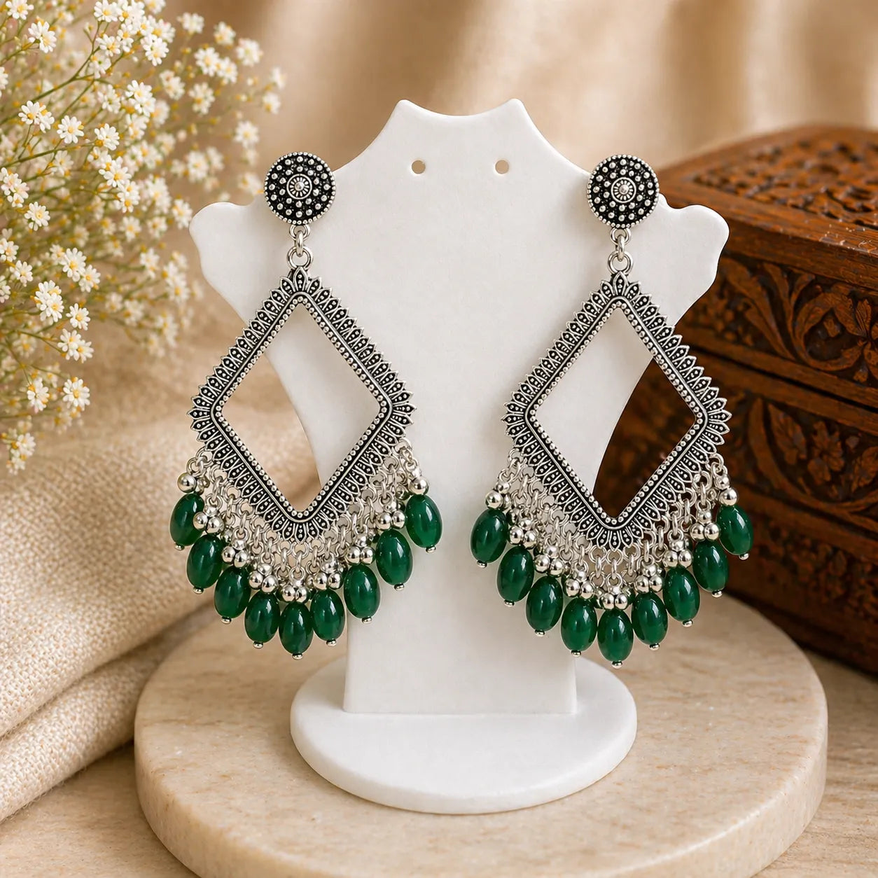 Ornate earrings with green beads on a white stand with a floral background
