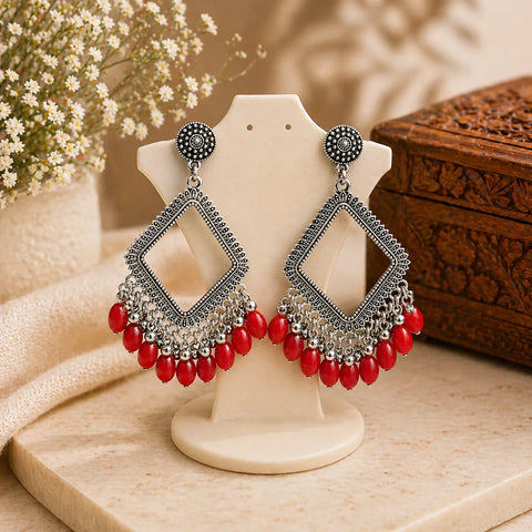 Ornate earrings with red beads on a display stand, with a wooden box and flowers in the background.