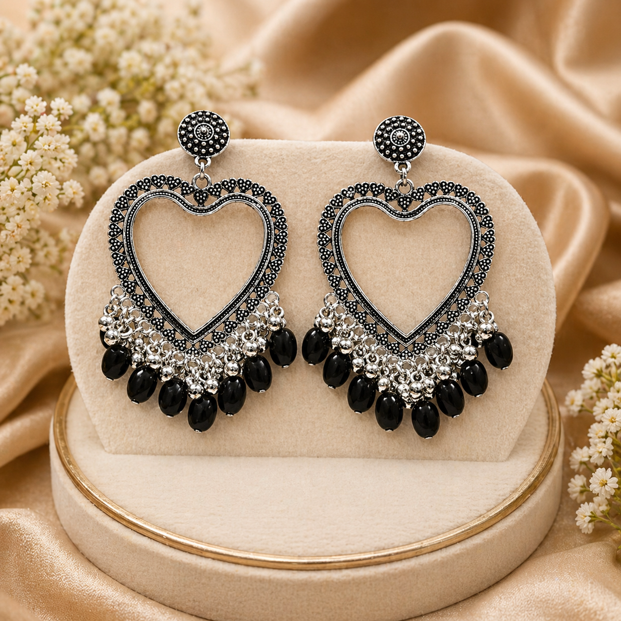 Heart-shaped earrings with black and silver details on a beige stand with flowers in the background
