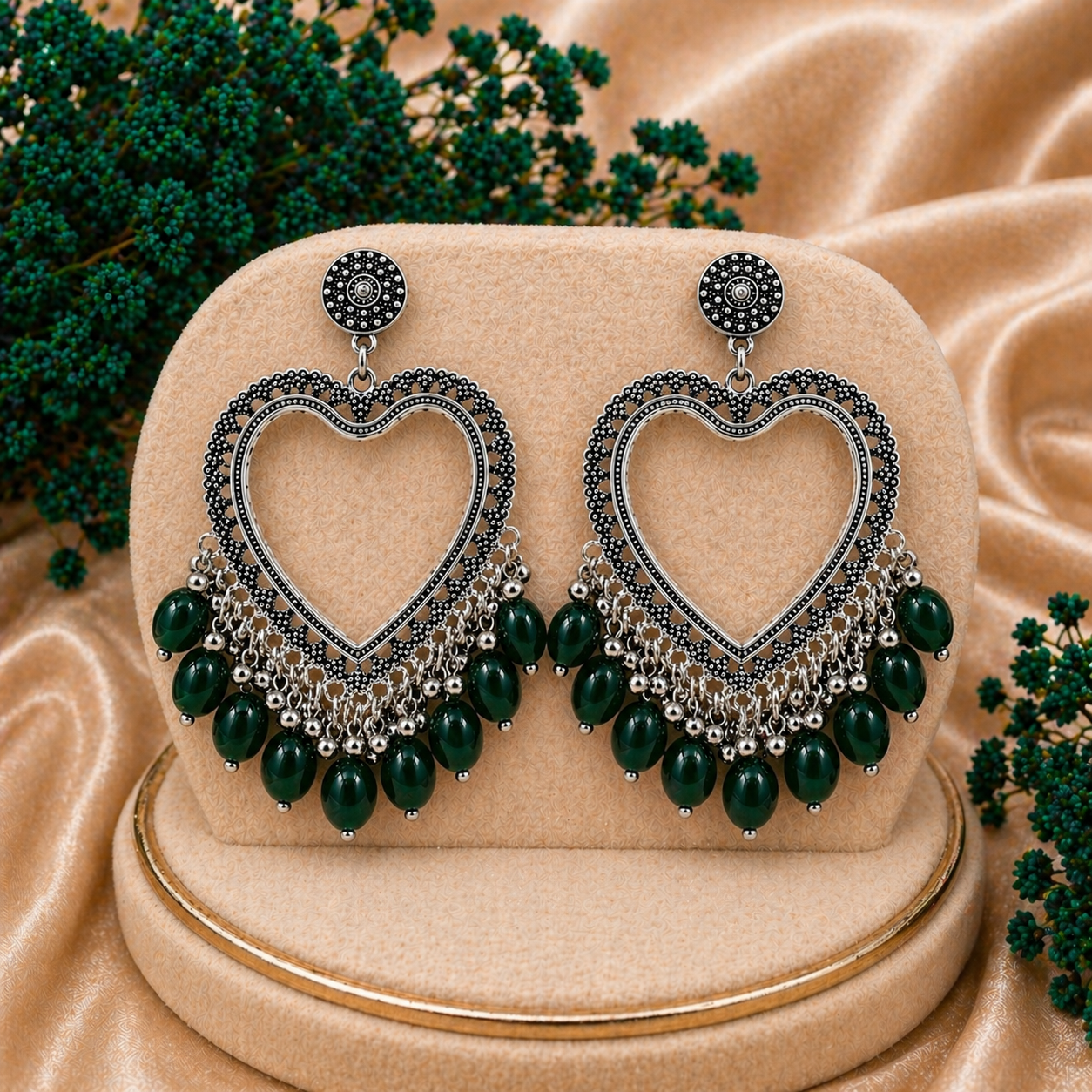 Heart-shaped earrings with green beads on a beige stand with green foliage in the background