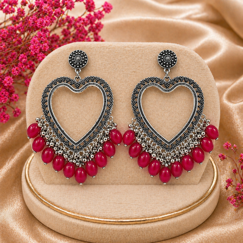 Heart-shaped earrings with red beads on a beige stand with pink flowers in the background