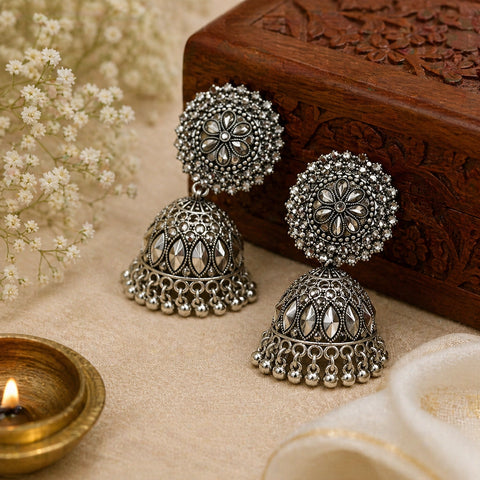 Pair of silver jhumka earrings on a textured surface with a wooden box and decorative elements.