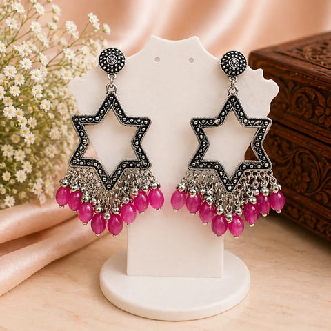Star-shaped earrings with pink beads on a white stand with a wooden box and flowers in the background.