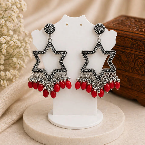 Star-shaped earrings with red beads on a white stand with a wooden box and flowers in the background.