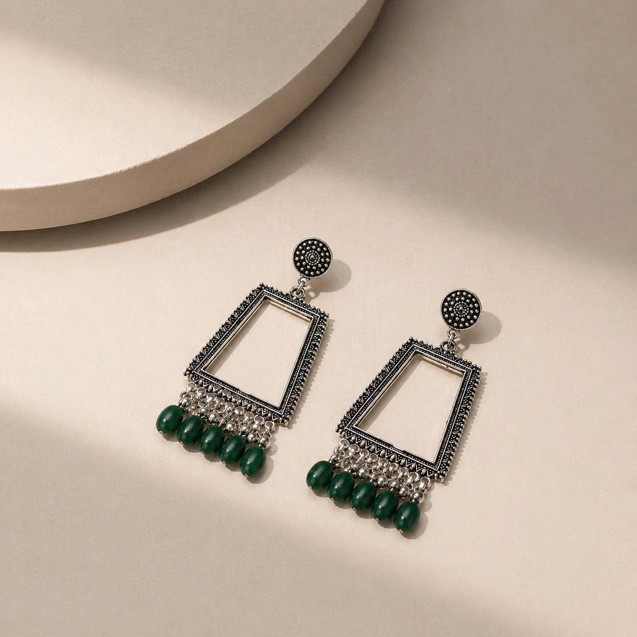 oxidised silver trapezium mirror drop earrings with green beads placed on beige surface top view jewellery image
