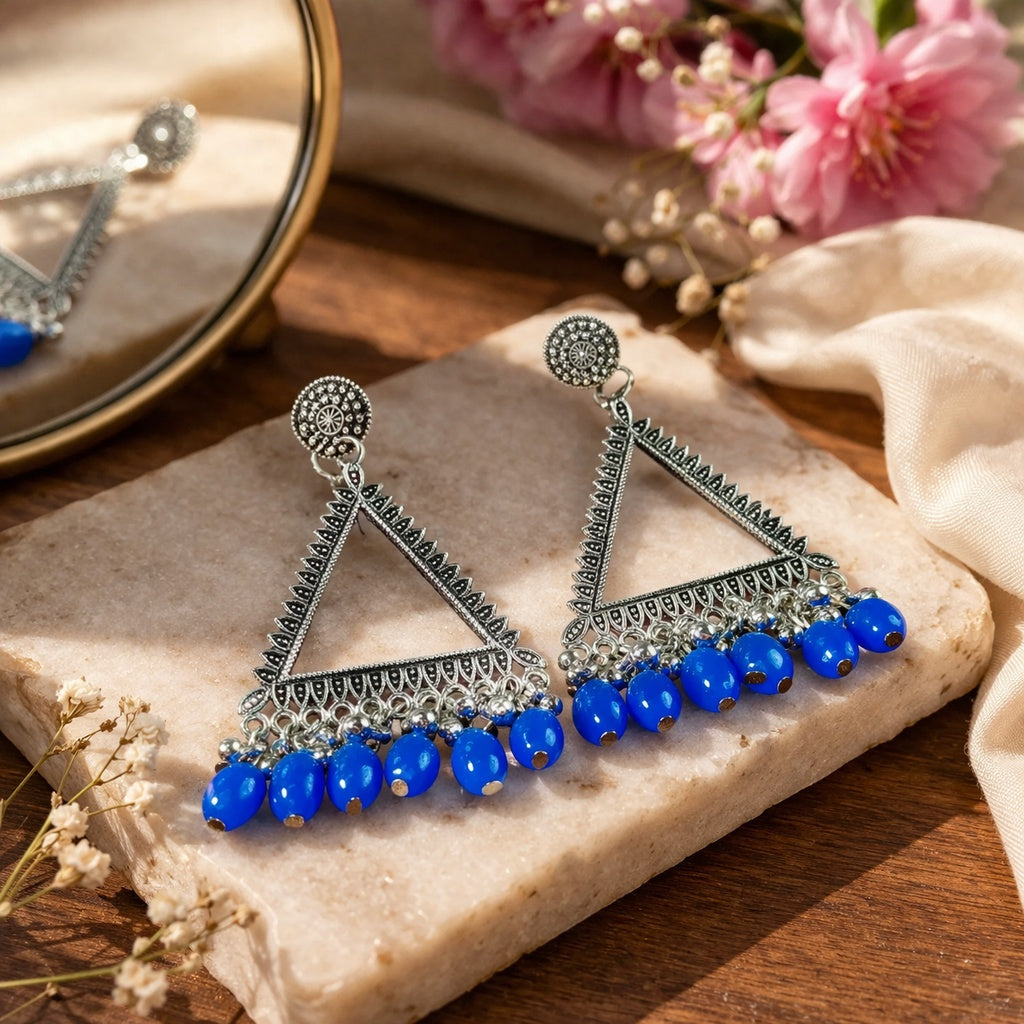 Triangular earrings with blue beads on a stone surface with flowers in the background