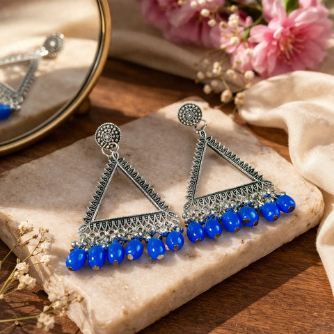 Triangular earrings with blue beads on a stone surface with flowers in the background