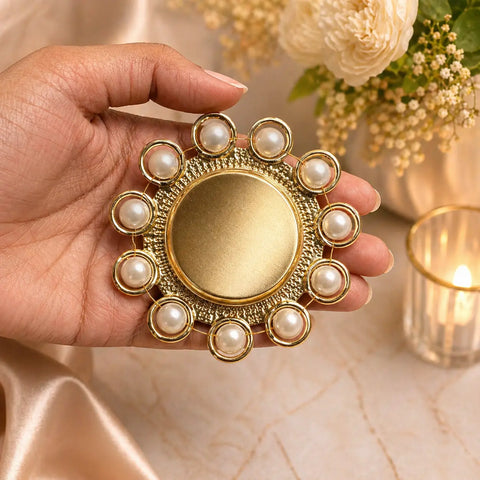 Gold decorative item with pearls held by a hand, with flowers and candle in the background