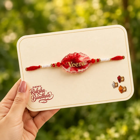 Personalized “Veera” handmade resin rakhi for brother with glossy red crystal resin centerpiece, white and red bead detailing, festive Raksha Bandhan card, and soft natural garden background.