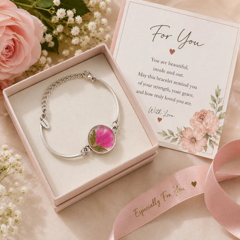 Silver bracelet with floral design in a pink box with a card, surrounded by flowers.