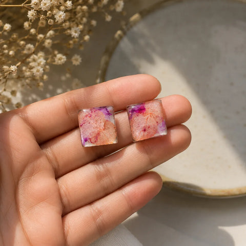 Handmade pink floral resin square stud earrings with glossy finish and dried flower detailing held in hand under soft natural sunlight