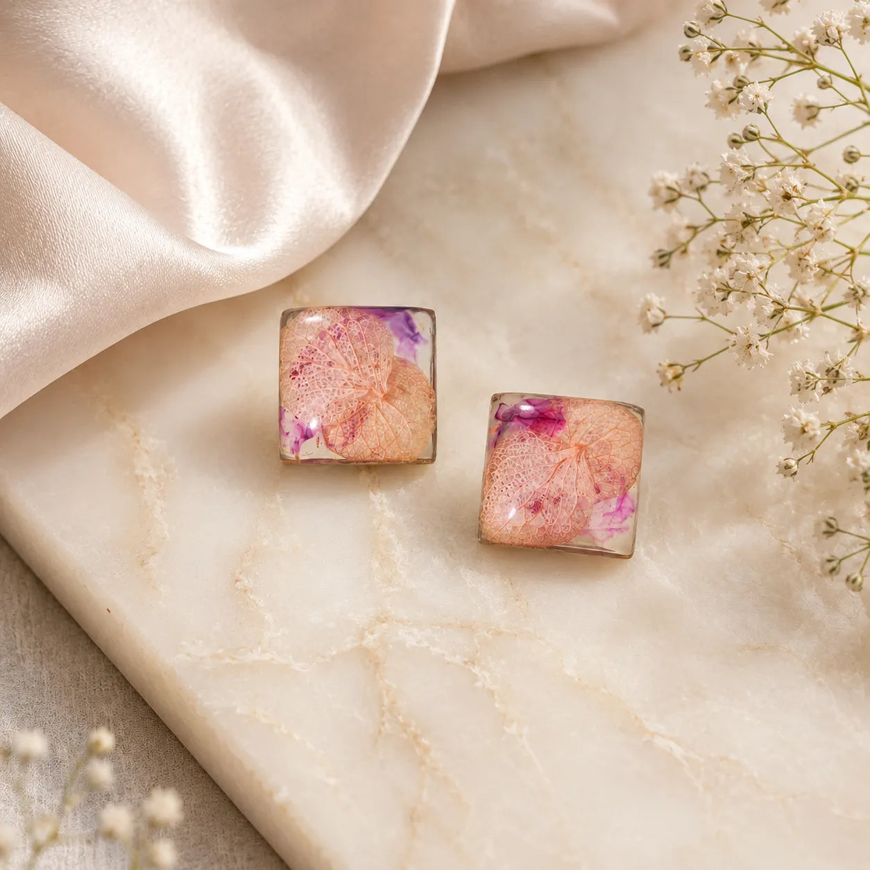 Top-view flat lay image of handmade pink floral resin square stud earrings placed on a marble surface with satin fabric and soft floral styling