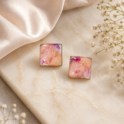 Top-view flat lay image of handmade pink floral resin square stud earrings placed on a marble surface with satin fabric and soft floral styling