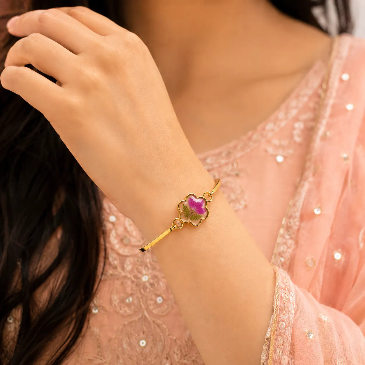 Close-up of a hand wearing a gold bracelet with a pink gemstone, against a blurred background.