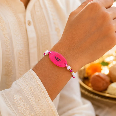 Hand wearing a pink bracelet with 'bhakt' on it, against a blurred background.