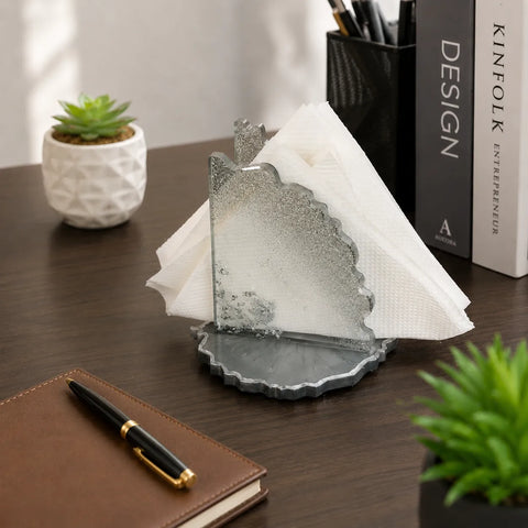 Decorative glass napkin holder on a desk with a pen, notebook, and books in the background.