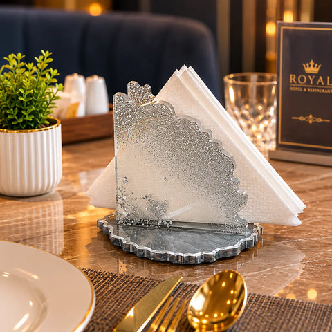 Decorative silver napkin holder on a table with cutlery and a plant.