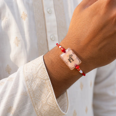 Bracelet with red beads and a clear rectangular charm on a wrist, against a light background.
