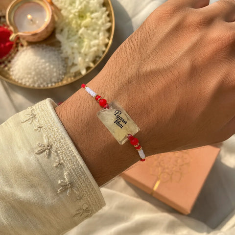 Hand wearing a red and white beaded bracelet with a clear tag, set against a soft focus background.