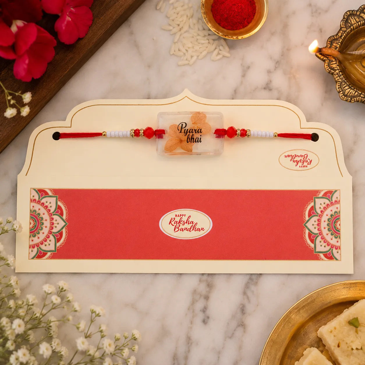 Rakhi with a card on a marble surface with flowers and sweets.
