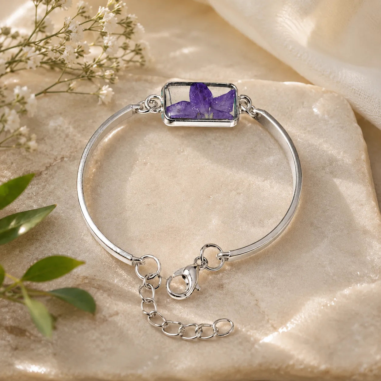 Silver bracelet with a purple gemstone on a beige stone surface with white flowers and green leaves.